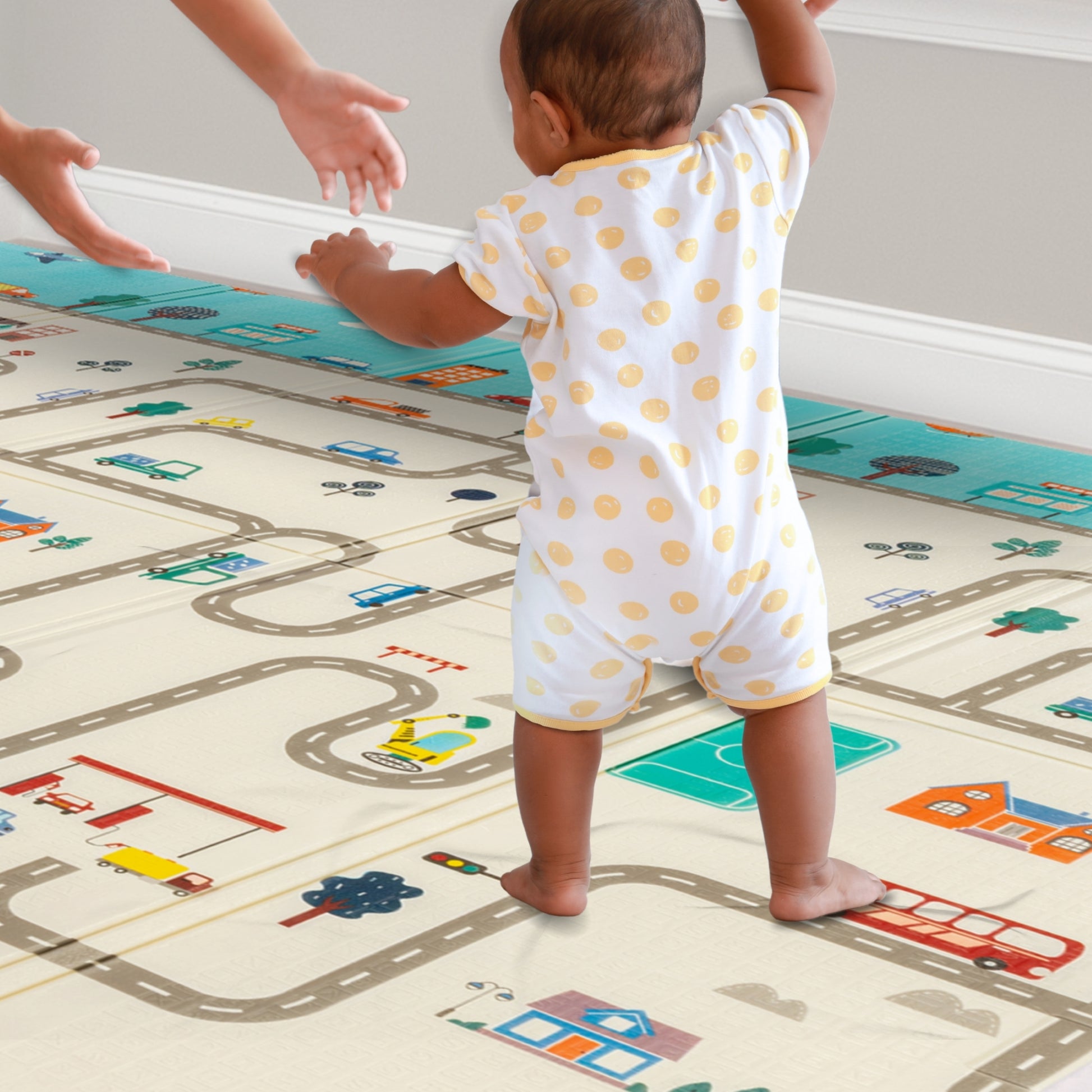 A baby taking first steps on a soft playmat with a parent holding their hands for support. The cushioned mat provides a safe, comfortable surface for the baby to walk on.