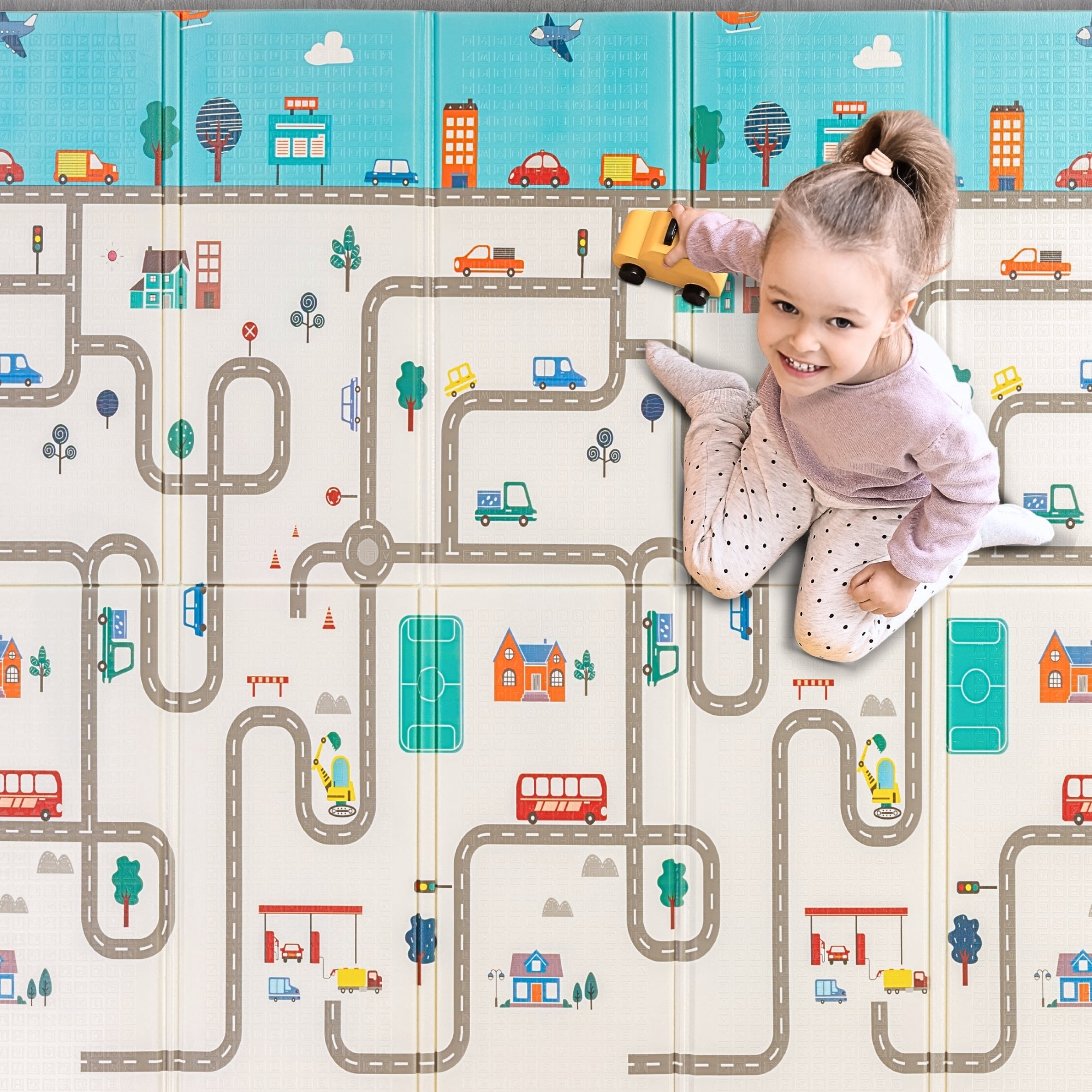 A toddler playing with a toy car on a city-themed playmat, driving along other vehicles pictures on colorful roads and buildings. The soft, cushioned mat features streets and landmarks for imaginative play.